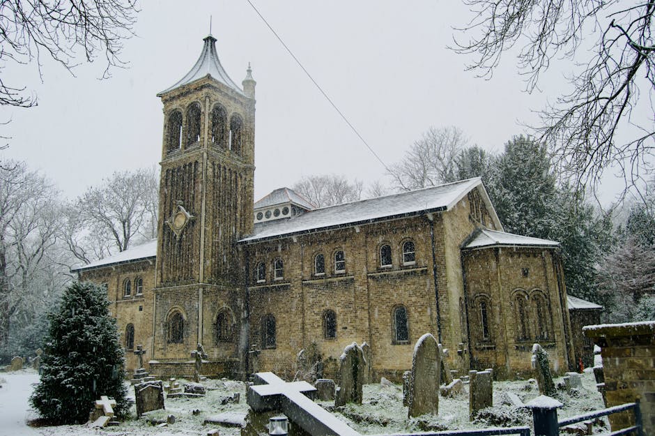 A historic stone church with a tall bell tower topped by a pointed roof, situated in a snowy landscape during winter. The church features arched windows and a weathered facade, with snow covering the 