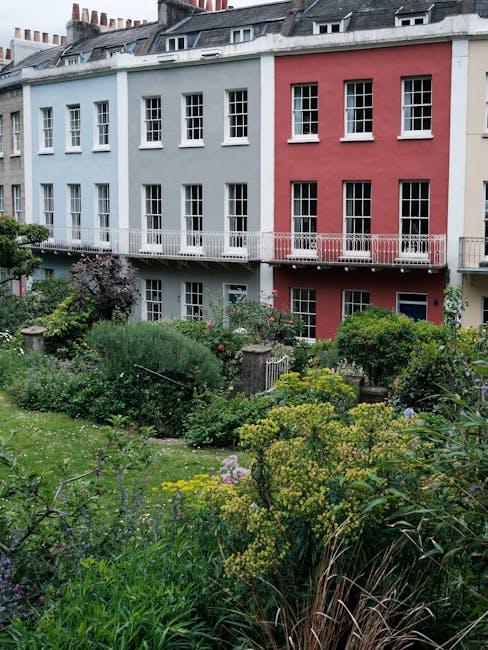 A colorful residential building with a light blue and pink façade features multiple white-framed sash windows and small balconies along a garden area. The garden showcases a variety of lush green shr