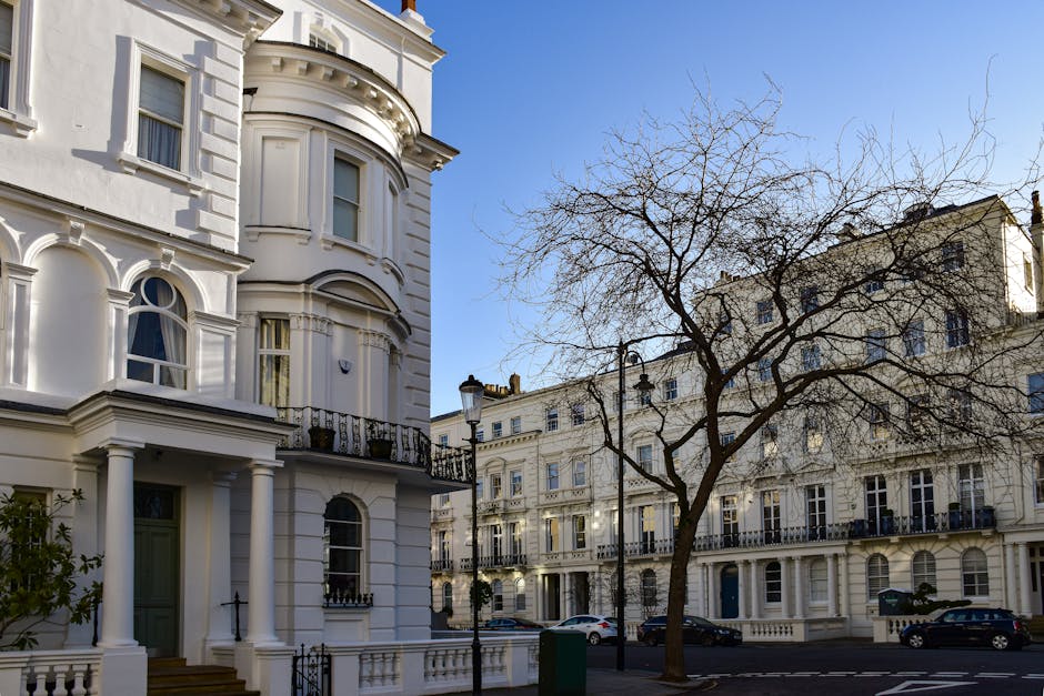 A colorful residential building with a light blue and pink façade features multiple white-framed sash windows and small balconies along a garden area. The garden showcases a variety of lush green shr